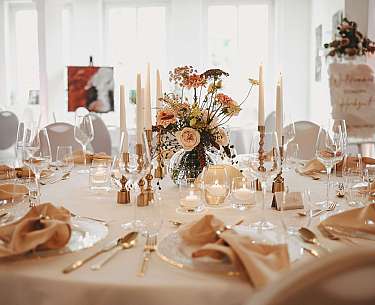 Festlich gedeckter runder Tisch mit Blumen, Kerzen und Stuhlhussen in hellem Raum.